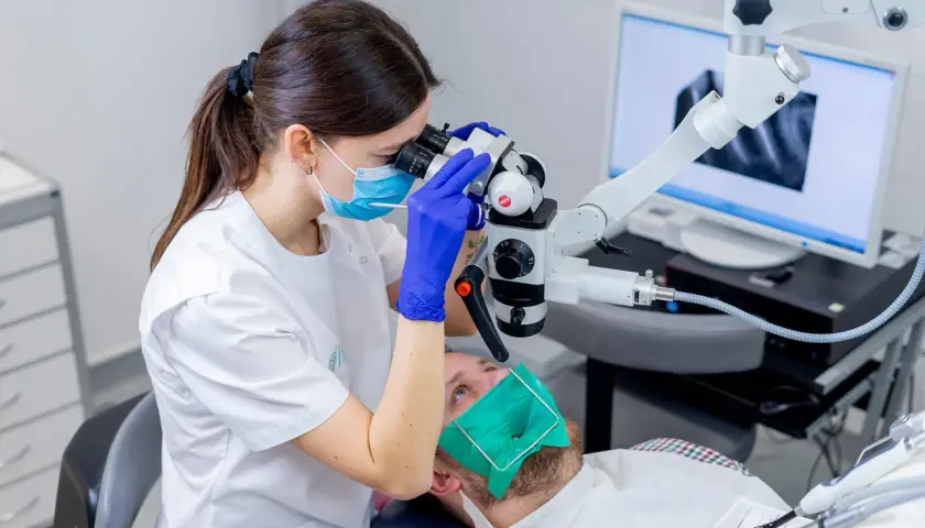 Dentystka w masce i rękawiczkach bada ząb pacjenta pod mikroskopem. Na ekranie widać zdjęcie rentgenowskie. To nowoczesne leczenie kanałowe, cena zabiegu zależy od skomplikowania.