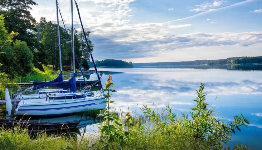 Żaglówki przy pomoście na spokojnym jeziorze. W tle las, a na wodzie odbijają się chmury. Mazury ciekawostki: idealne miejsce na relaks i żeglowanie.