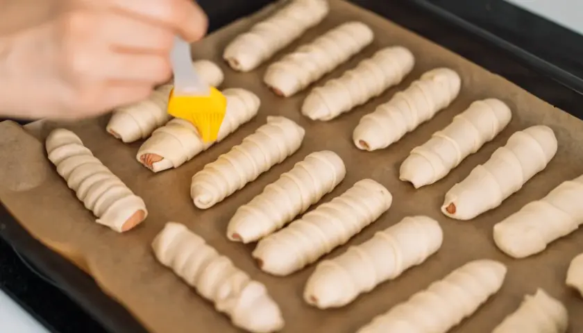 Ręka smaruje pędzelkiem parówki zawinięte w ciasto, przygotowując je do pieczenia. To prosty przepis na parowance.