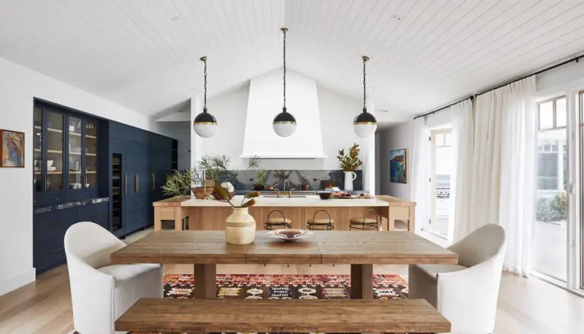 A modern lake house interior featuring a rustic wooden dining table, plush white chairs, and a sleek navy kitchen island.