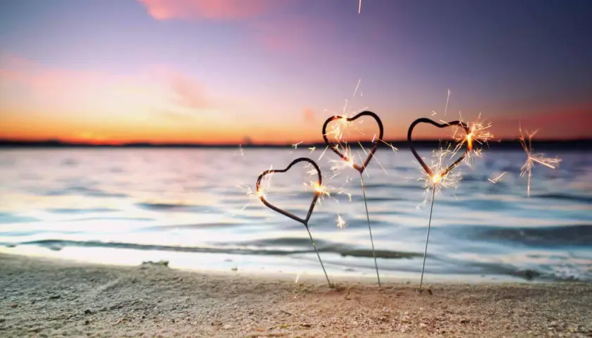 Drei Herz-Wunderkerzen funkeln am Strand. Ein romantischer Silvester am Meer mit einem farbenprächtigen Himmel.