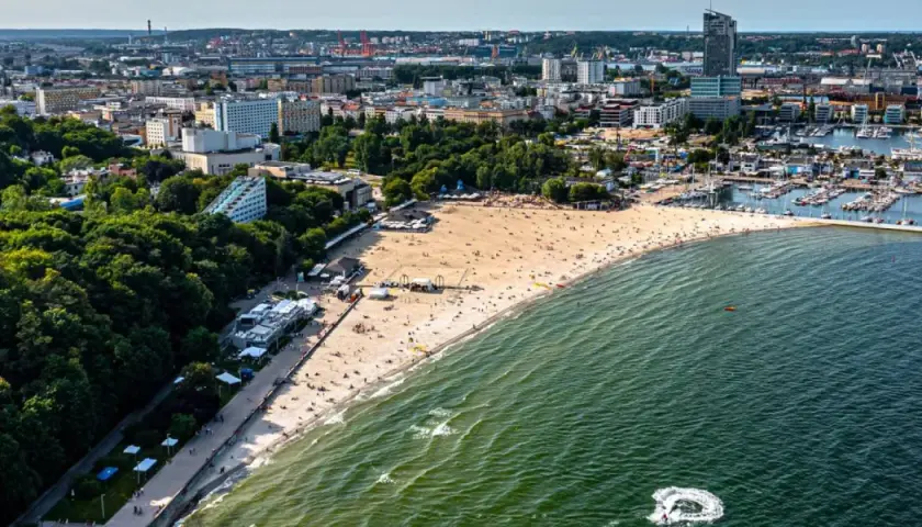 Słoneczny bulwar nadmorski Gdynia tętni życiem. Plaża pełna ludzi, a w oddali port z żaglówkami i miejska panorama.