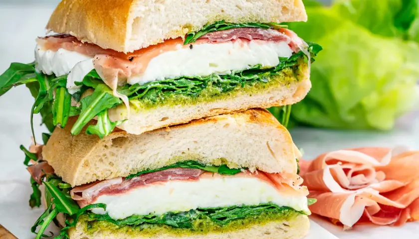 Dos bocadillos italianos apilados, rellenos de mozzarella fresca, prosciutto, rúcula y pesto, sobre pan rústico.