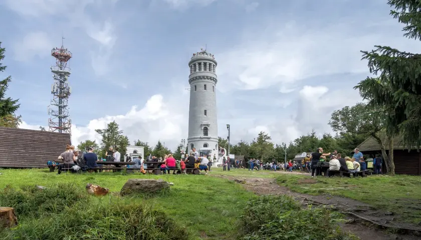Ludzie odpoczywają przy wieży widokowej i maszcie telekomunikacyjnym. W oddali widać wielką sowę na tle chmur.