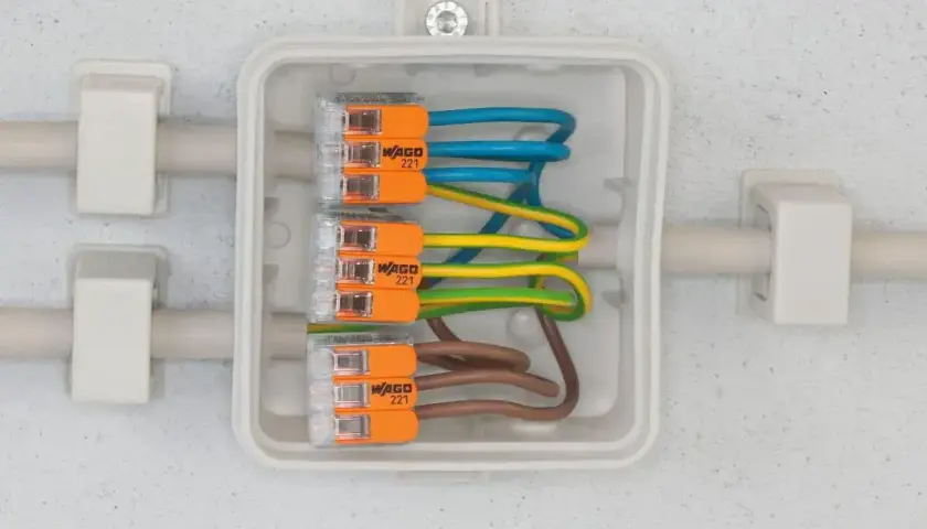 Bezpieczne przyłącze elektryczne z przewodami w kolorach niebieskim, żółto-zielonym i brązowym, połączonymi złączkami WAGO 221 w białej puszce.