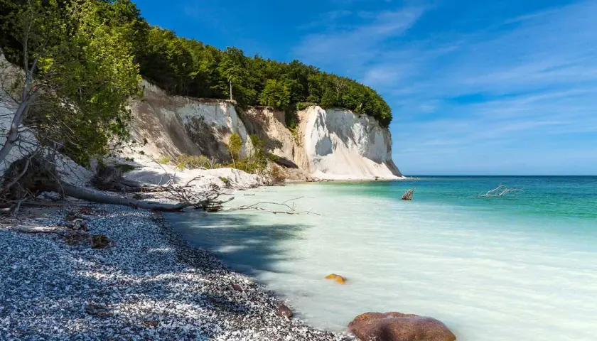 Weiße Kreidefelsen ragen steil auf, bewaldet von oben. Die Ostsee bei Sassnitz schimmert türkisblau, ein Kiesstrand lädt zum Verweilen ein.