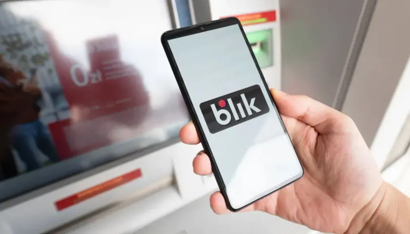 Dłoń trzyma smartfon z logo Blik przed bankomatem. Blik działa w Polsce i kilku innych krajach Europy.