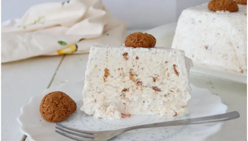 Una fetta di semifreddo agli amaretti, guarnita con un amaretto intero e un altro sul dolce intero sullo sfondo.