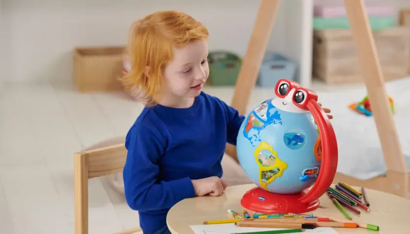 Un bambino di 5 anni con capelli rossi esplora il mondo con un globo interattivo, circondato da matite colorate.