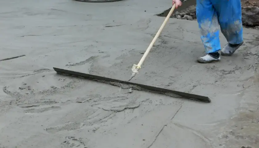Pracownik w niebieskich ogrodniczkach wygładza świeży beton długą pacą. Trwa wygrzewanie posadzki.