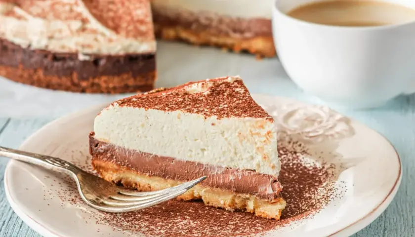 Pyszna tarta z mascarpone i czekoladą, posypana kakao, czeka na widelczyk. Obok filiżanka kawy.