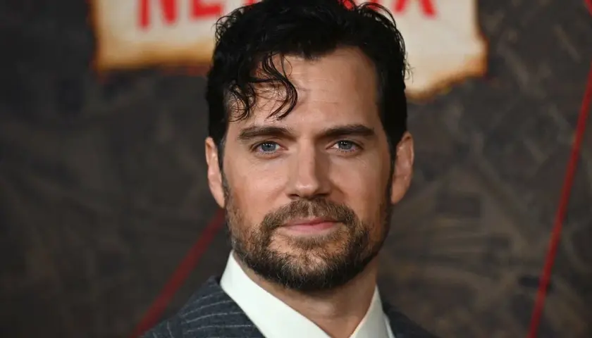 Henry Cavill w garniturze i czerwonym krawacie, z zarostem i niebieskimi oczami.