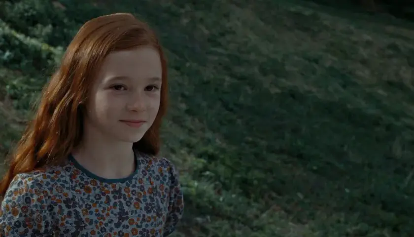 Młoda Lily Evans, przyszła matka Harrego Pottera, z rudymi włosami i w kwiecistej sukience, uśmiecha się na tle zielonego wzgórza.