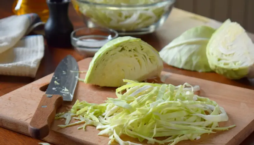 Frisch geschnittener Weißkohl liegt auf einem Holzbrett, bereit für ein Sauerkraut Rezept. Daneben ein Messer und Schüssel mit Kohl.