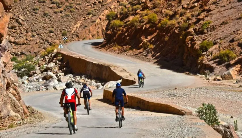 Ciclistas disfrutan de una aventura de **Marruecos en bici** por un camino sinuoso entre montañas rocosas y vegetación escasa.