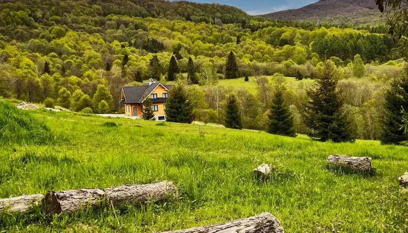 Zielony, pagórkowaty krajobraz Bieszczad z drewnianym domkiem. Idealne miejsce, by dowiedzieć się, jak dojechać w Bieszczady pociągiem i cieszyć się naturą.