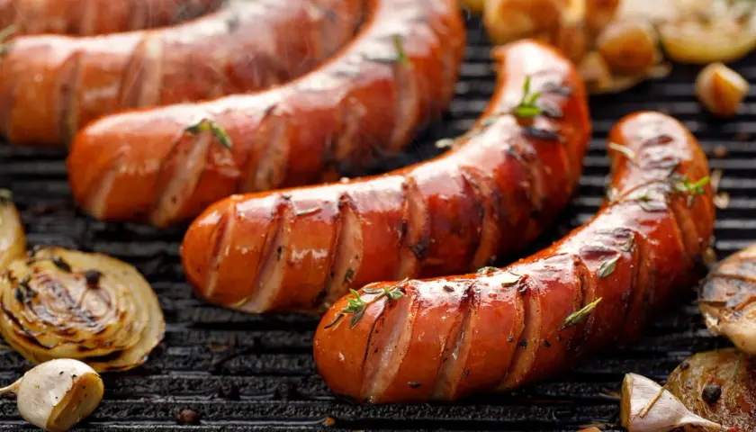 Soczysta kiełbasa z grilla, parująca i pachnąca ziołami. Obok grillowana cebula i czosnek. Idealna na letnie wieczory, choć warto pamiętać o jej kcal.