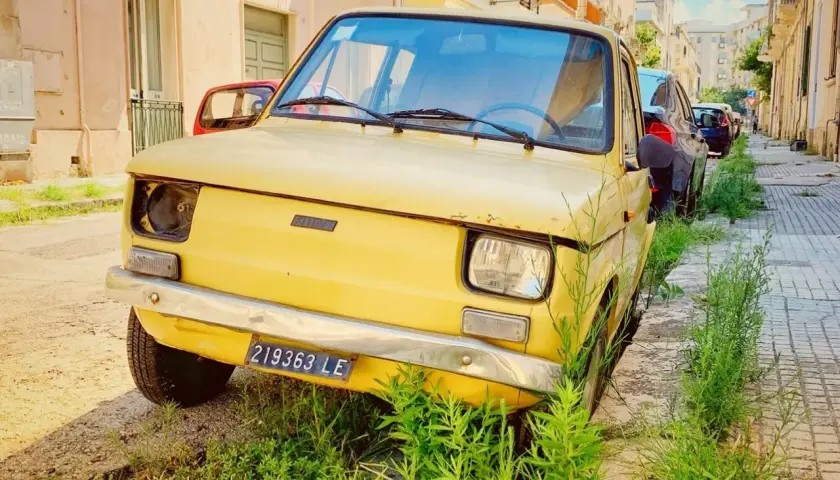 A vintage yellow Fiat, a symbol of driving in Puglia, sits parked on a sun-drenched street, overgrown with lush green grass.