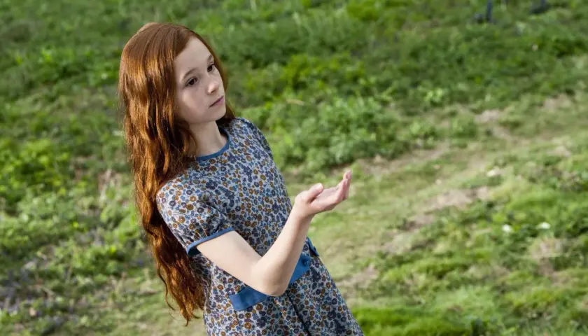 Ruda dziewczynka w kwiecistej sukience, Lily Potter, wyciąga dłoń w stronę zielonej łąki.
