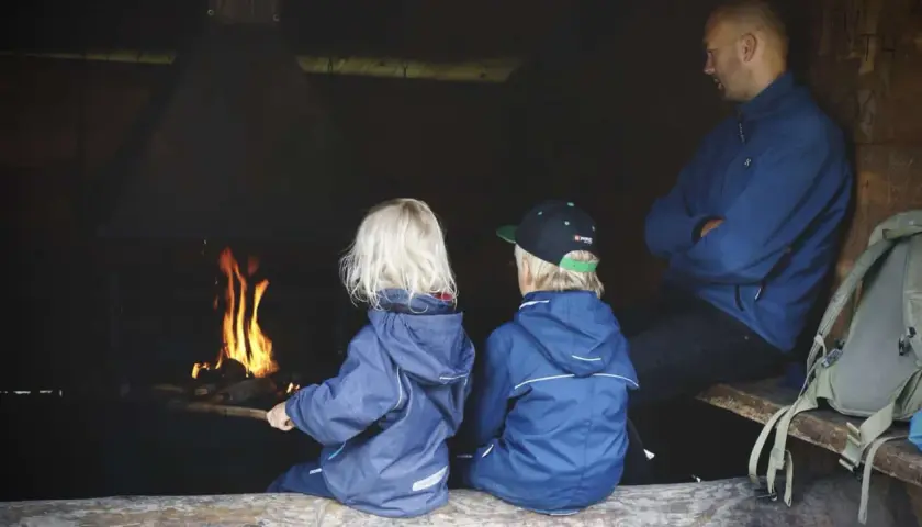 Två barn och en vuxen sitter vid en eld. En mysig paus under vandring med barn.