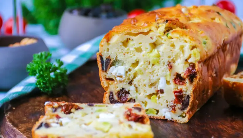Un délicieux cake salé aux olives, tomates séchées et feta, coupé en tranches sur une planche en bois.