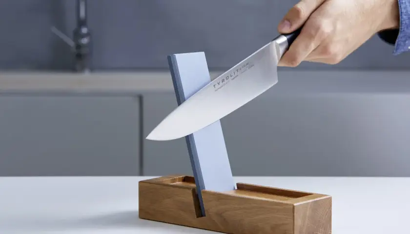 Ein Messer wird auf einem Schleifstein geschärft, der in einem Holzständer steckt. Dieser Schleifstein ist ein wichtiger Helfer, um die Schneide scharf zu halten, fast wie ein **dichtungsschaber** für die Klinge.