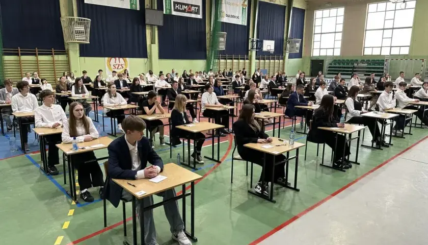 Uczniowie siedzą przy ławkach w sali gimnastycznej podczas egzaminu.