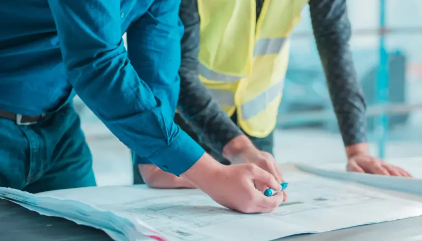 Deux hommes examinent des plans. L'un, en chemise bleue, pointe un détail avec un stylo. L'autre, en gilet jaune, pose ses mains sur les documents. Un constat huissier avant travaux est en cours.