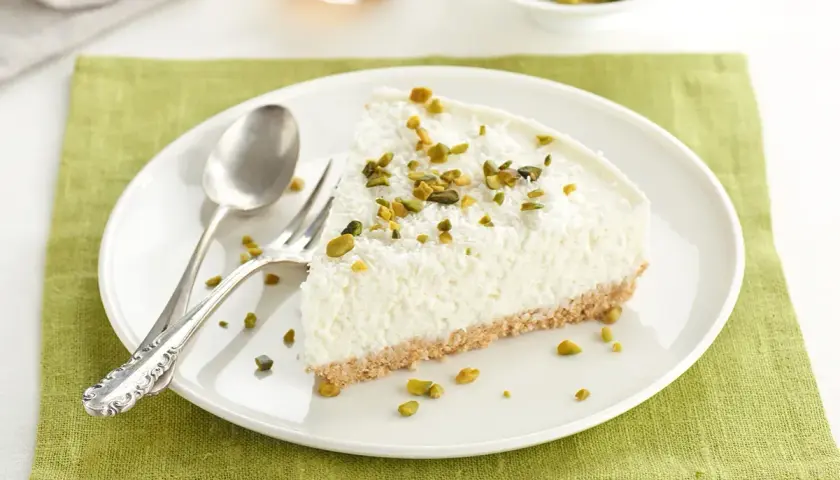 Rebanada de tarta de coco mousse, cubierta con coco rallado y pistachos picados, sobre plato blanco con cubiertos.