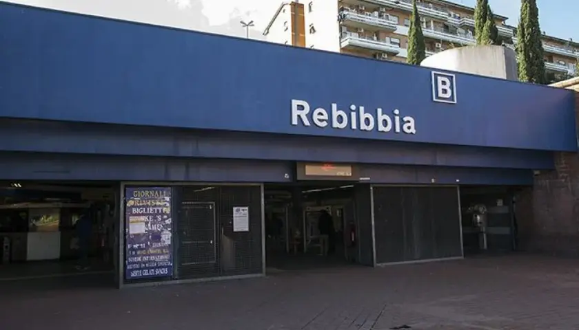 Ingresso della metro Rebibbia a Roma, con insegna blu e lettere bianche. Edifici residenziali sullo sfondo.