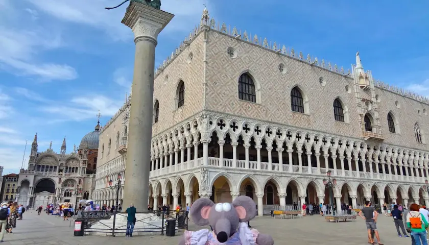 Une souris en robe rose devant l'entrée du Palais des Doges à Venise, avec la colonne de Saint-Marc et la basilique Saint-Marc en arrière-plan.