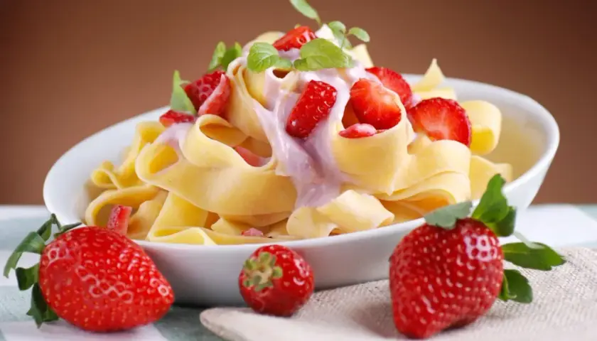 Pappardelle z kremowym sosem i kawałkami truskawek, ozdobione listkami mięty. Obok talerza leżą świeże truskawki.