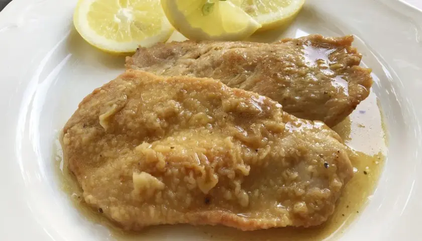 Deliziose scaloppine al limone, succulente e dorate, servite con spicchi di limone fresco su un piatto bianco.
