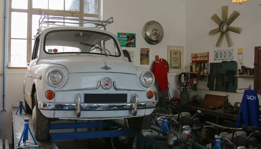 Biały Fiat 600 na podnośniku w warsztacie, gotowy do napraw. W tle narzędzia i motocykle, a nad nimi lampa zapewniająca oświetlenie kanału samochodowego.