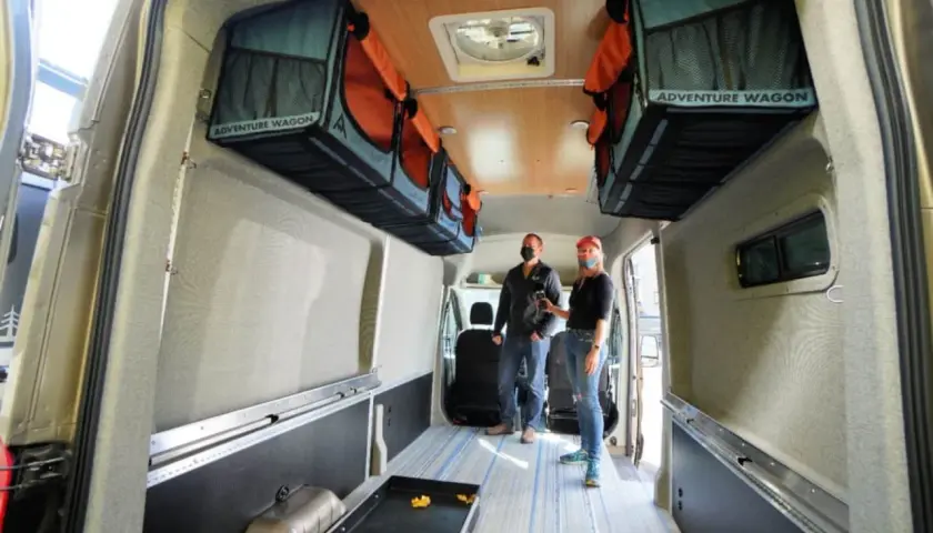 Aménagement fourgon par un professionnel : deux personnes admirent l'intérieur aménagé avec des rangements suspendus "Adventure Wagon" et un sol à rayures bleues.