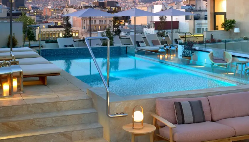 Piscina en la azotea del Barceló Raval con vistas a la ciudad al atardecer. Tumbonas, sofás y luces crean un ambiente acogedor.