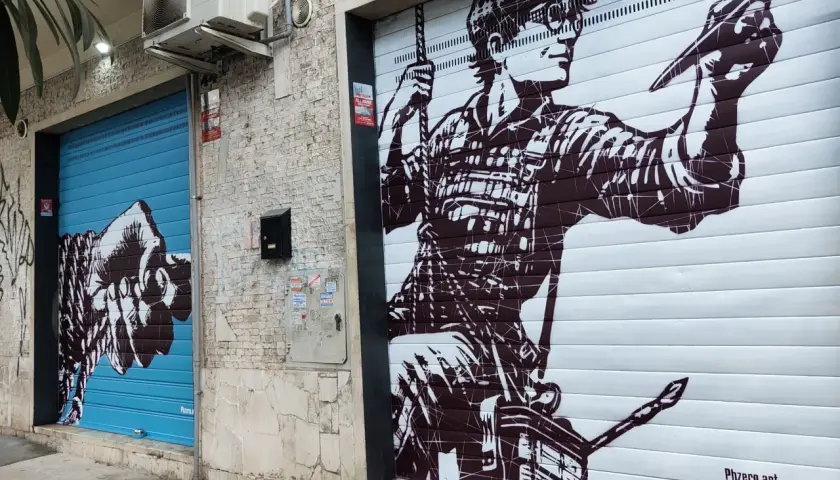 Un pugno su saracinesca blu e un artista al lavoro su un altro saracinesca: arte urbana in un quartiere murales di Roma.