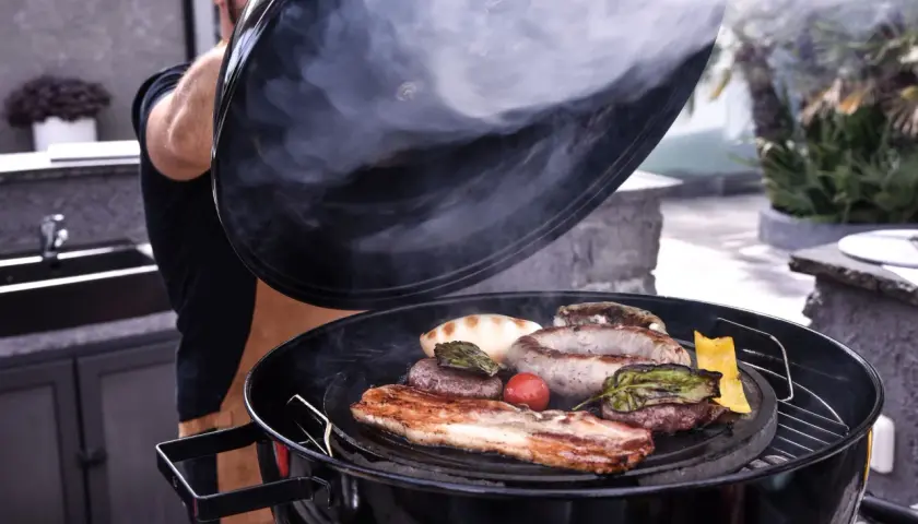 Un homme soulève le couvercle d'un barbecue fumant, révélant une sélection alléchante de viande au barbecue : saucisses, steaks, lard et légumes grillés.