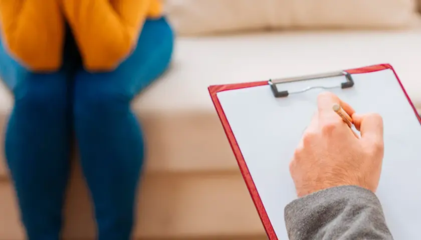 Seduta in terapia, con un professionista che prende appunti. Un passo verso il benessere, forse un giorno con assicurazione psicologi obbligatoria.