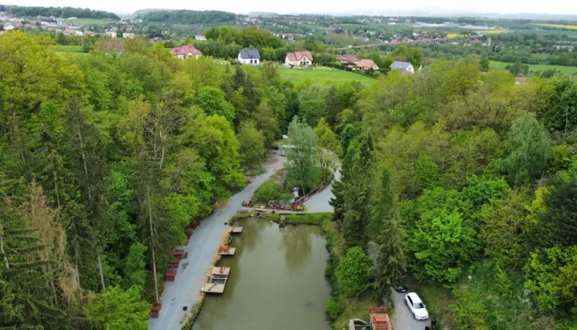 Widok z lotu ptaka na łowisko pstrągowe otoczone zielonymi drzewami, z domami w tle.