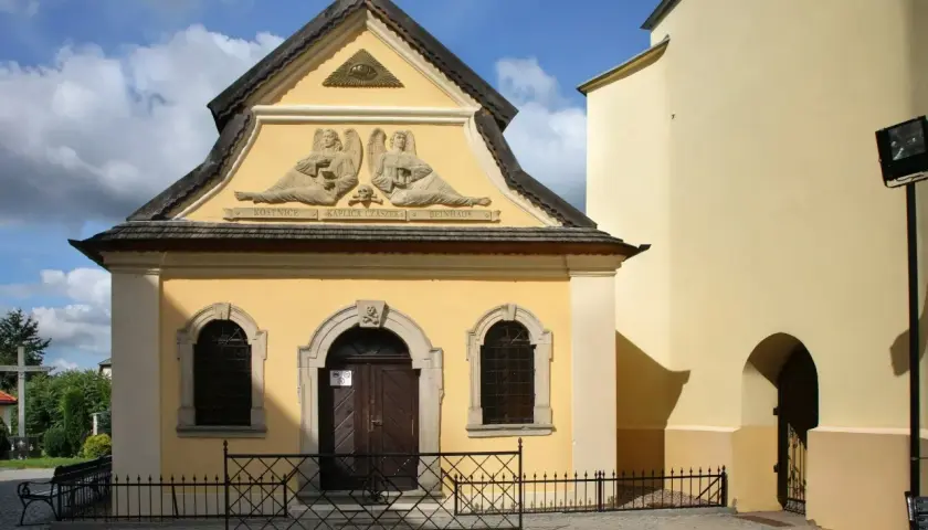 Żółta fasada kaplicy czaszek z płaskorzeźbami aniołów i symbolem oka w trójkącie. Pod nimi napis "Kostnice Kaplica Czaszek".