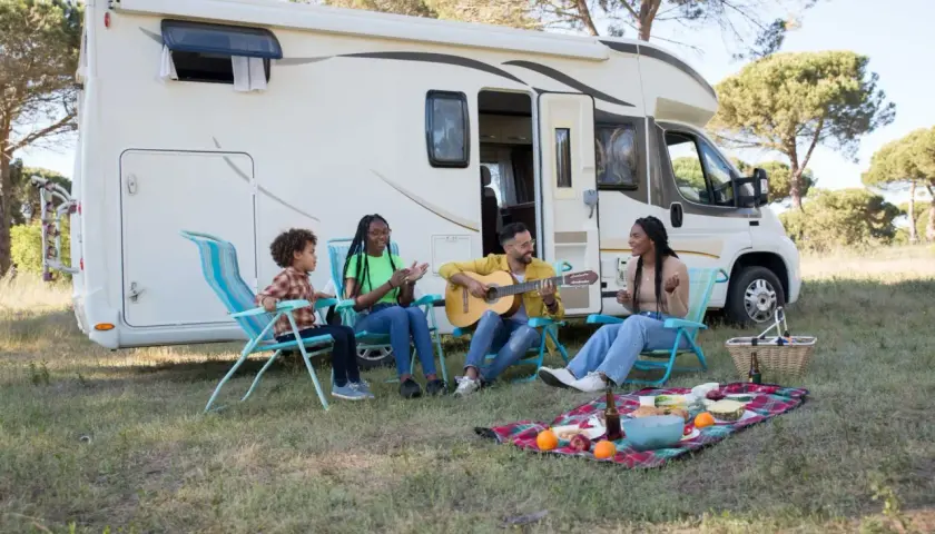 Familie genießt das Zelten mit Kindern. Ein Mann spielt Gitarre, während die anderen singen und lachen. Ein Picknickkorb und Obst liegen auf einer Decke.