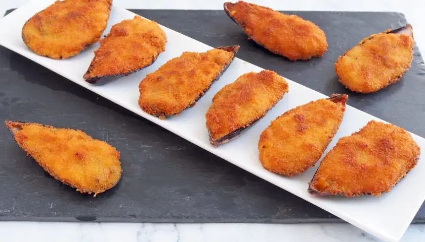 Ocho mejillones tigre dorados y crujientes, servidos en una bandeja blanca sobre una pizarra.