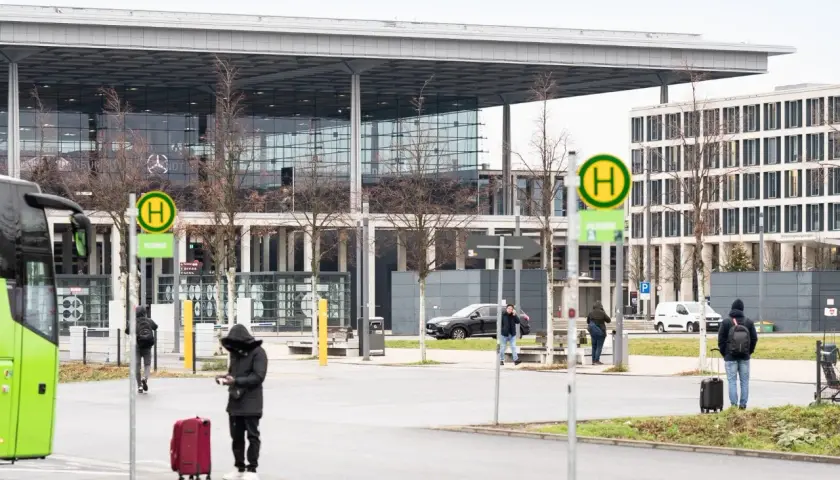 Przystanek autobusowy z zielonym autobusem, ludźmi z bagażami i nowoczesnym budynkiem w tle.