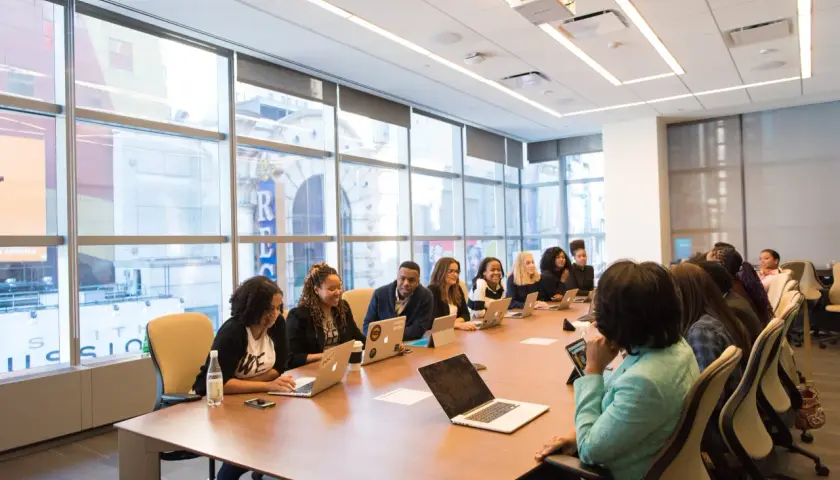 A diverse group of professionals engage in government leadership training, collaborating around a conference table with laptops and tablets.