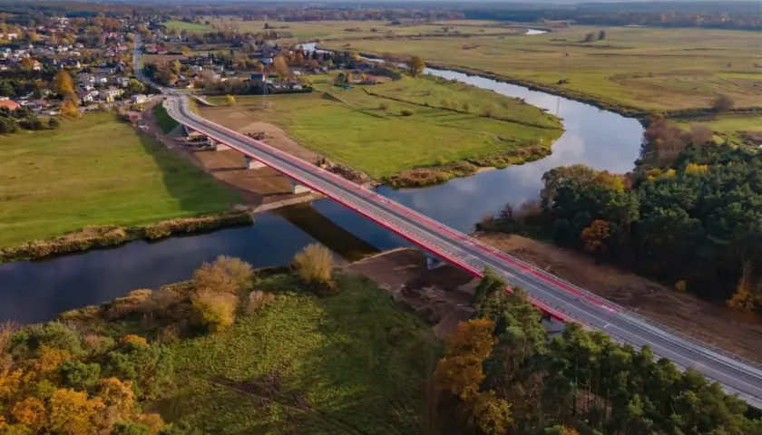 Nowy, czerwony most na warcie przecina spokojną rzekę, łącząc malowniczą wieś z zielonymi polami i lasami jesienią.