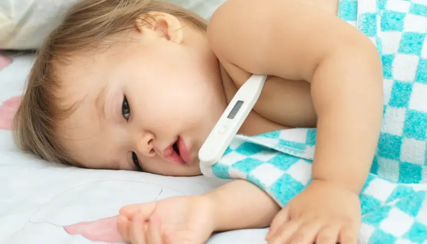 Bambino con termometro sotto l'ascella, avvolto in un asciugamano a quadri. La febbre che non scende bambini è una preoccupazione.