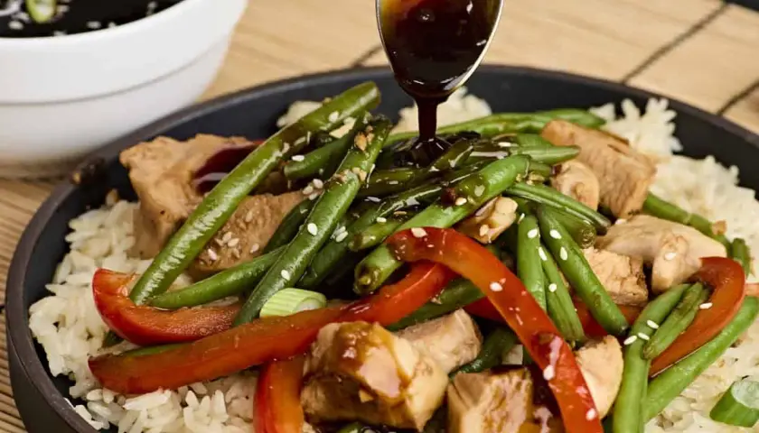 A spoon drizzles teriyaki sauce over a dish of rice, chicken, green beans, and red peppers, garnished with sesame seeds.