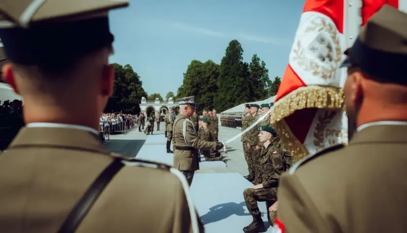 Podczas uroczystości wojskowej, oficer wręcza szablę klęczącemu podporucznikowi. W tle stoją żołnierze i widzowie.