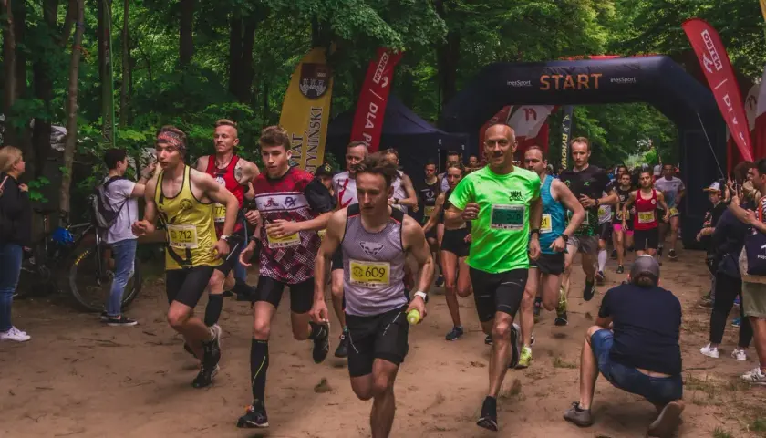 Uczestnicy biegu ultra trail startują w lesie. Widać łuk startowy, flagi i biegaczy w kolorowych koszulkach.